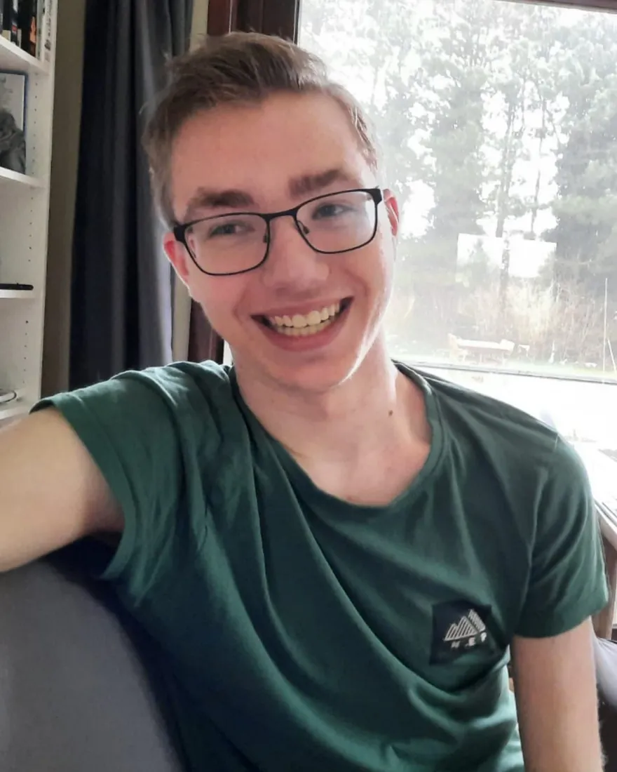 Niek lacht in de camera, in een groen T-shirt, thuis bij het raam