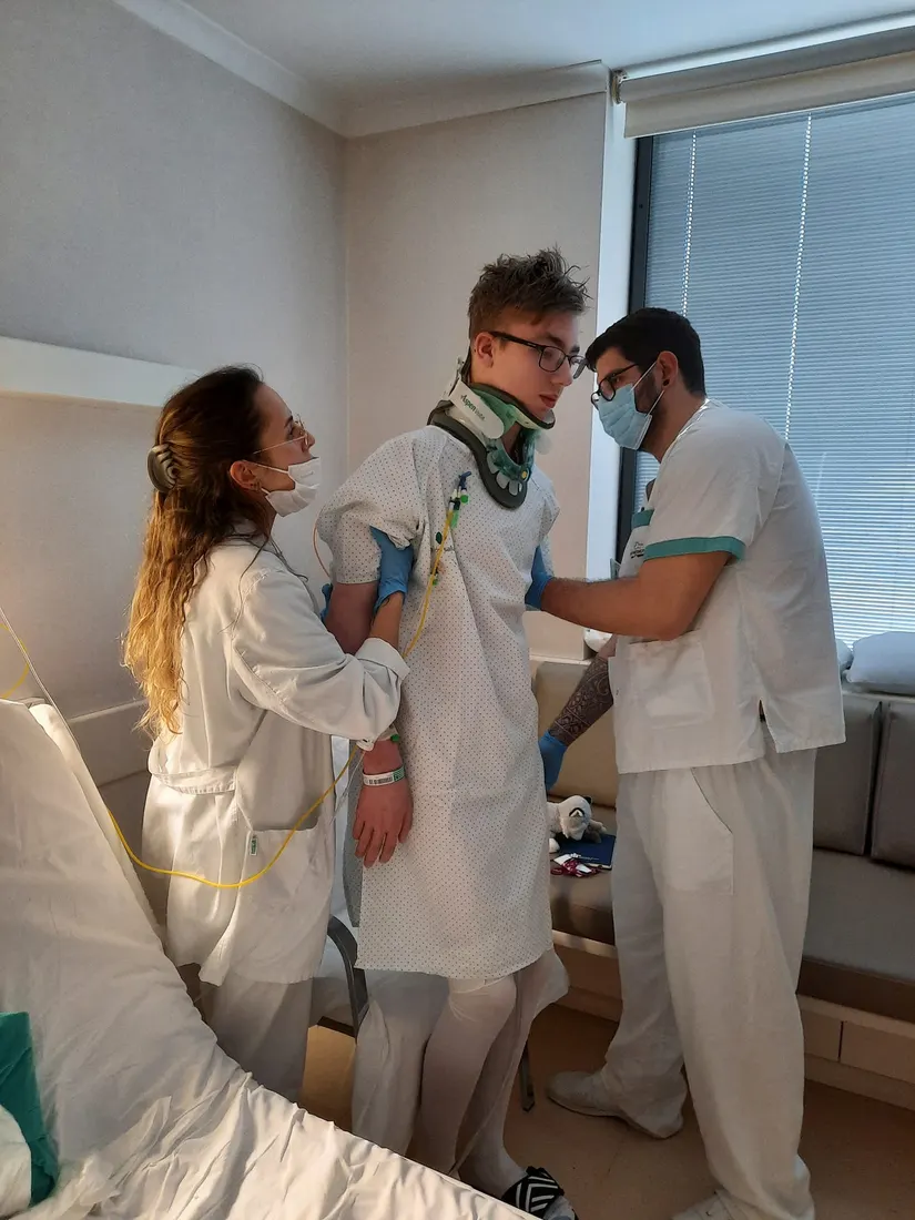 Niek staat met nekkraag tussen twee zorgverleners in zijn ziekenhuiskamer